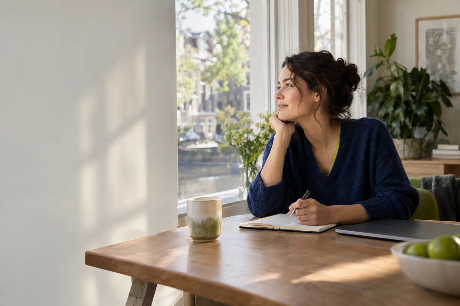 Een vrouw in een donkerblauwe trui zit aan een houten tafel bij het raam met een opengeslagen notitieboek en kijkt glimlachend naar buiten