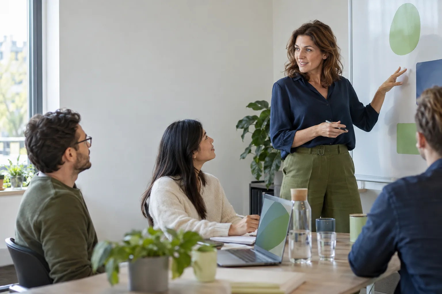 Een vrouw staat aan het hoofd van een tafel en praat met twee collega's, die naar haar opkijken