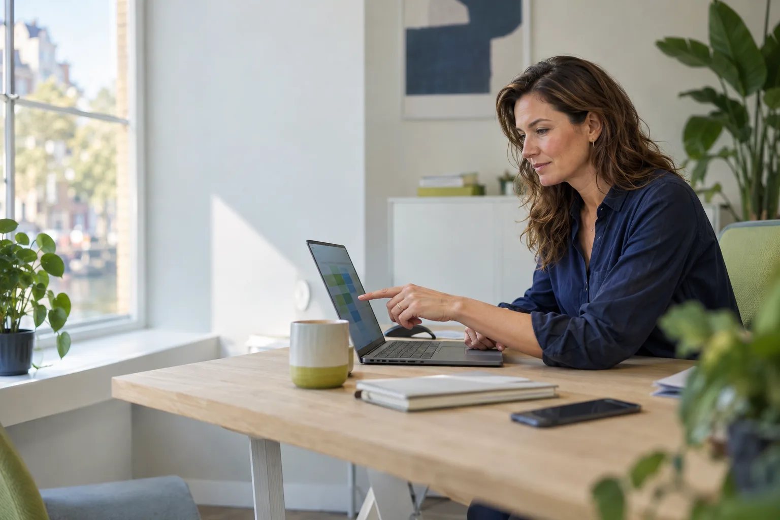 Een vrouw in een donkerblauwe blouse wijst aan een houten bureau in een lichte kamer iets aan op het scherm van haar laptop, met een groene mok naast zich