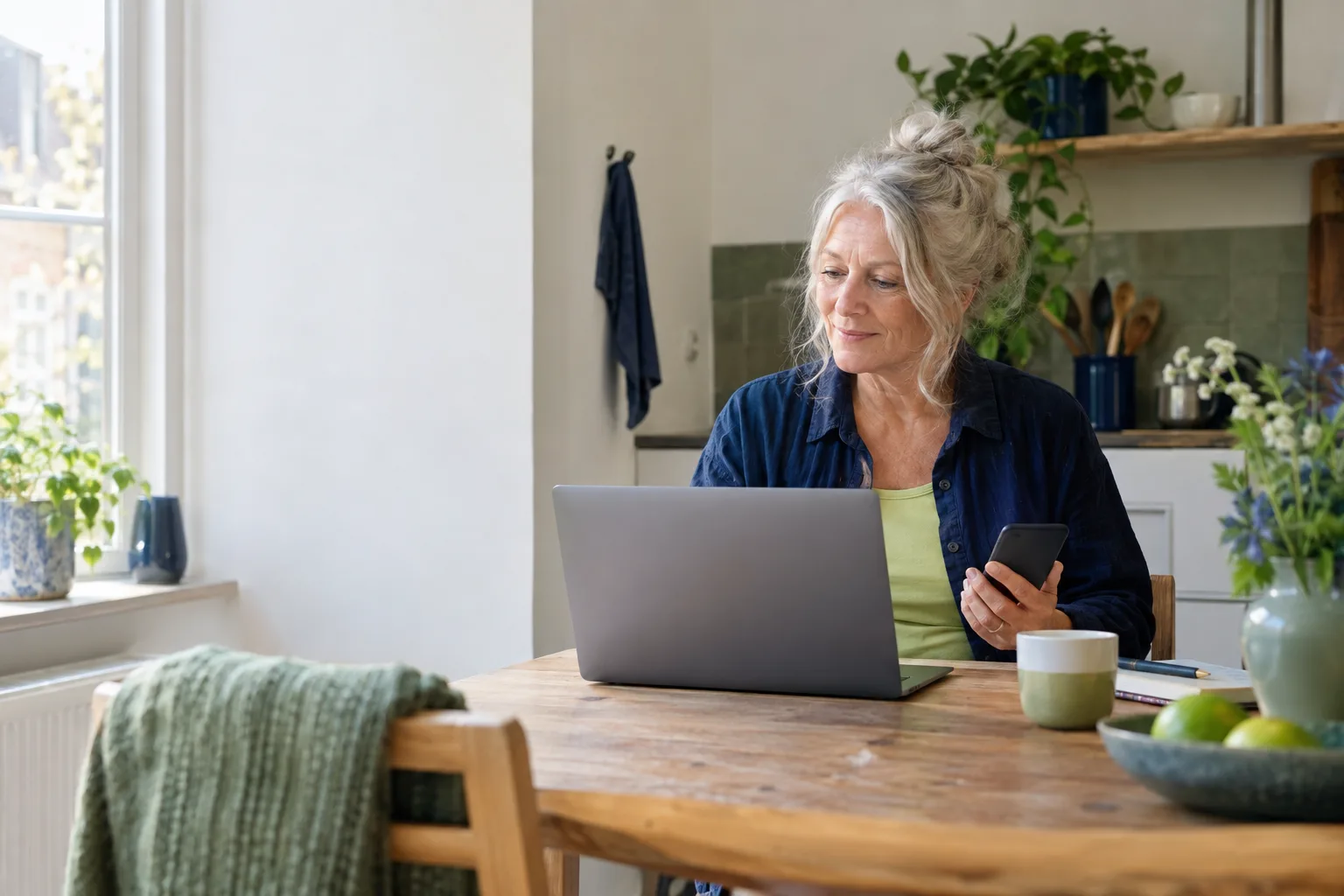 Een vrouw met grijs haar werkt aan een laptop en houdt haar telefoon in de hand