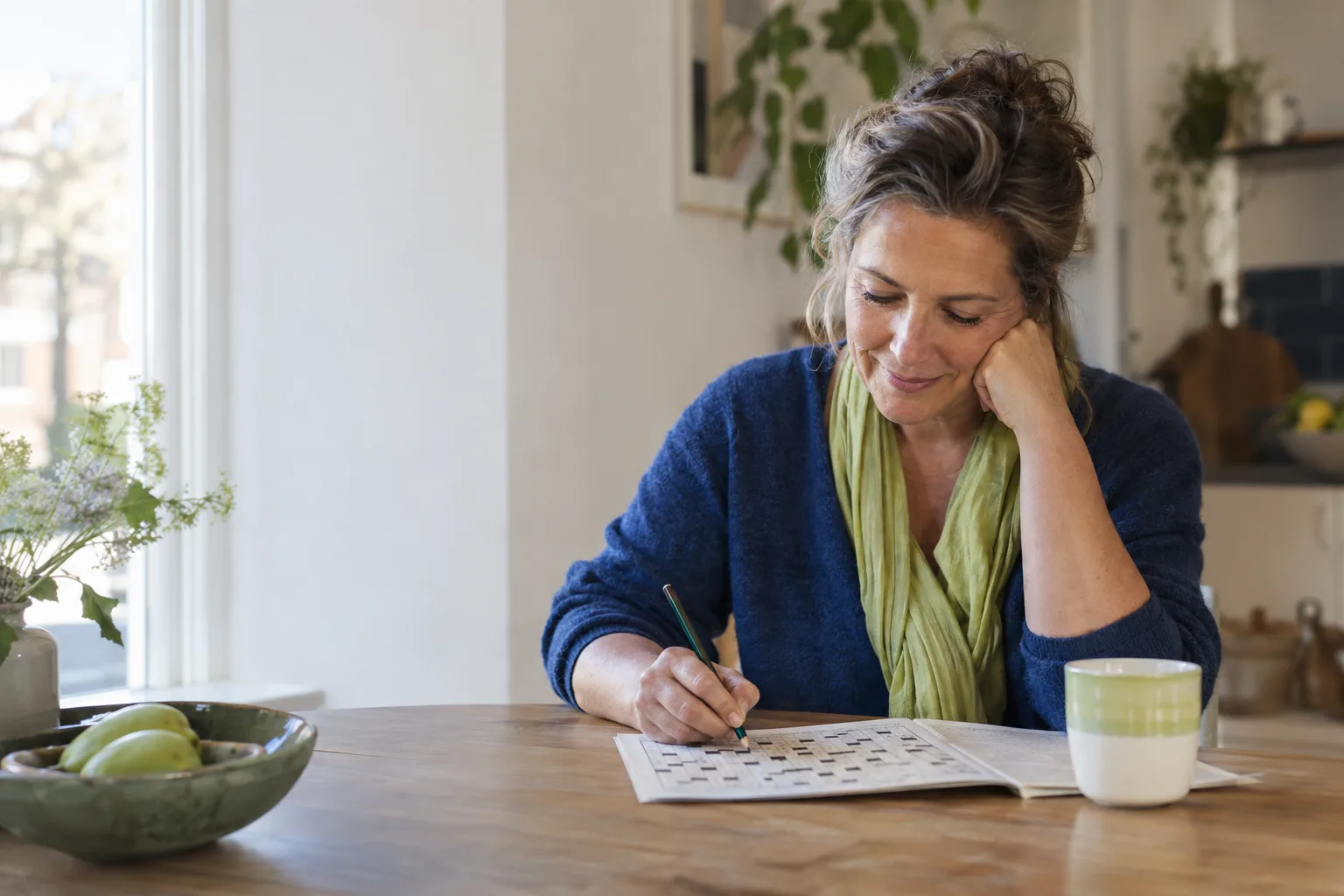 Een vrouw zit aan een keukentafel over een puzzel gebogen, met een potlood in haar hand